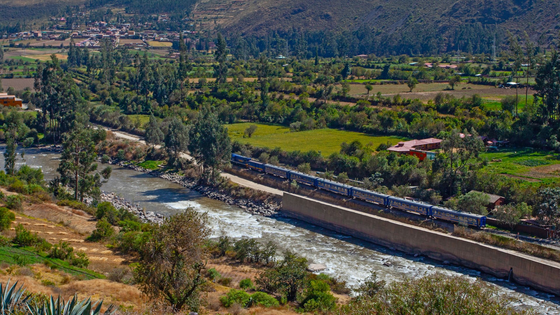 Panoramic view of a blue luxury train traversing a green valley alongside a river, representing the scenic travel on a 1 Day Luxury Tour to Machu Picchu - Aremika Travel
