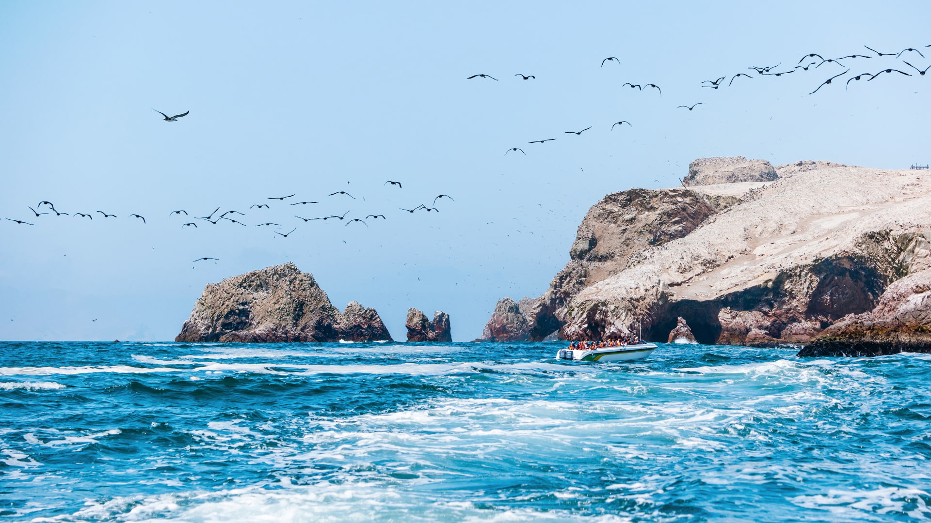 Tourist boat navigating near guano-covered rock formations with hundreds of birds flying over Ballestas Islands during a Tour Paracas and Huacachina Full Day - Aremika Travel