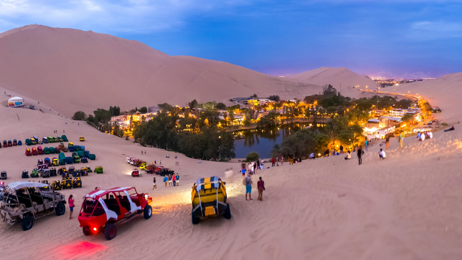 Panoramic view of Huacachina Oasis at dusk with dune buggies and people on surrounding sand dunes, part of a Tour Paracas and Huacachina Full Day - Aremika Travel
